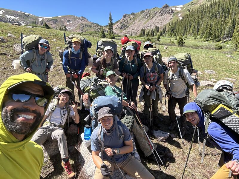 A group of hikers are posing for a photo in a mountainous area. They are wearing backpacks and hiking gear. The weather appears to be sunny. The hikers are smiling and appear to be enjoying their time in nature. Some of them are wearing hats and sunglasses. The background includes mountains, trees, and grass.